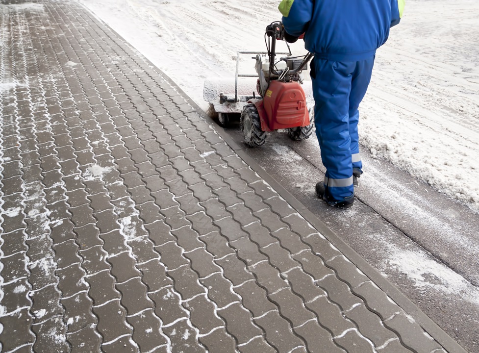 153760291 Person in blue winter clothes uses a snow removal machine on a snowy, icy sidewalk.