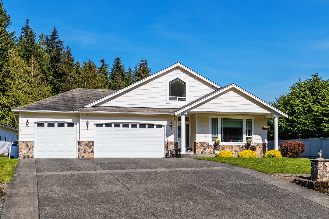Single Story Suburban Home Exterior with Large Driveway White single-story house with three-car garage and wide driveway, surrounded by trees and shrubs.