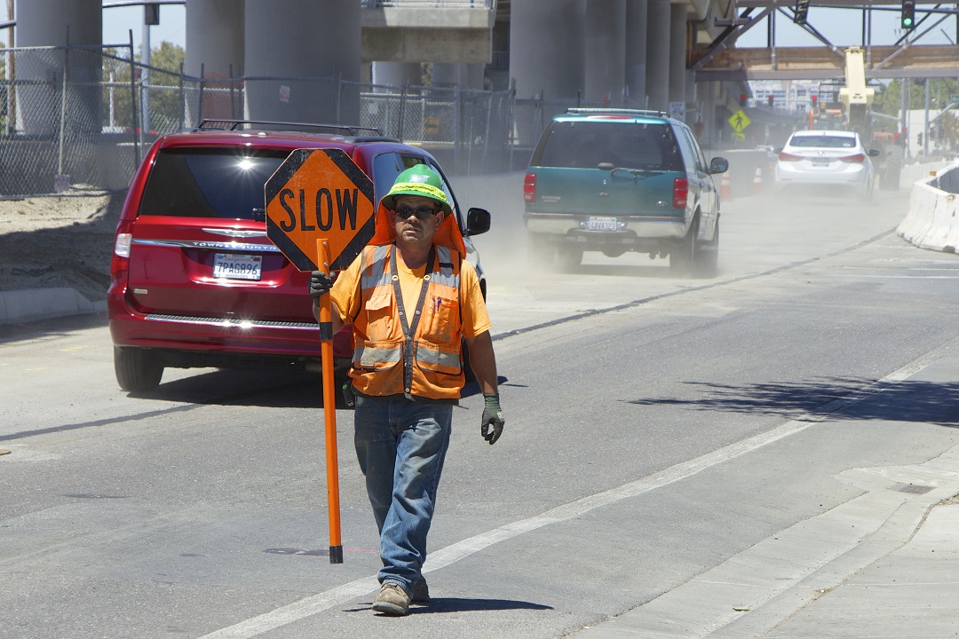 Construction worker in safety gear holds a "SLOW" sign as cars drive by on a dusty road.