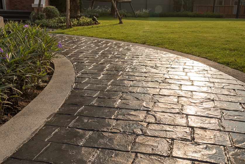 Stamp concrete texture pattern and background. Curved stone pathway bordered by grass and plants, with sunlight reflecting off the surface.