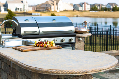 Outdoor kitchen with a grill, sink, and kebabs on a cutting board, overlooking a pond and houses.