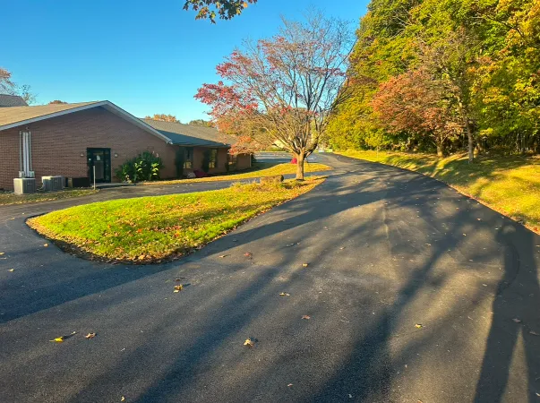 A paved road curves past a brick building and trees with autumn leaves under a clear blue sky.