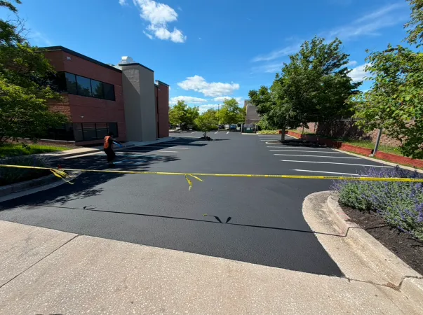 Freshly paved parking lot blocked off with yellow tape, next to a brick building and trees on a sunny day.