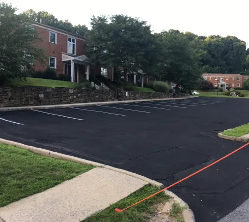 Empty, freshly paved parking lot with white lines, next to brick buildings and green trees.