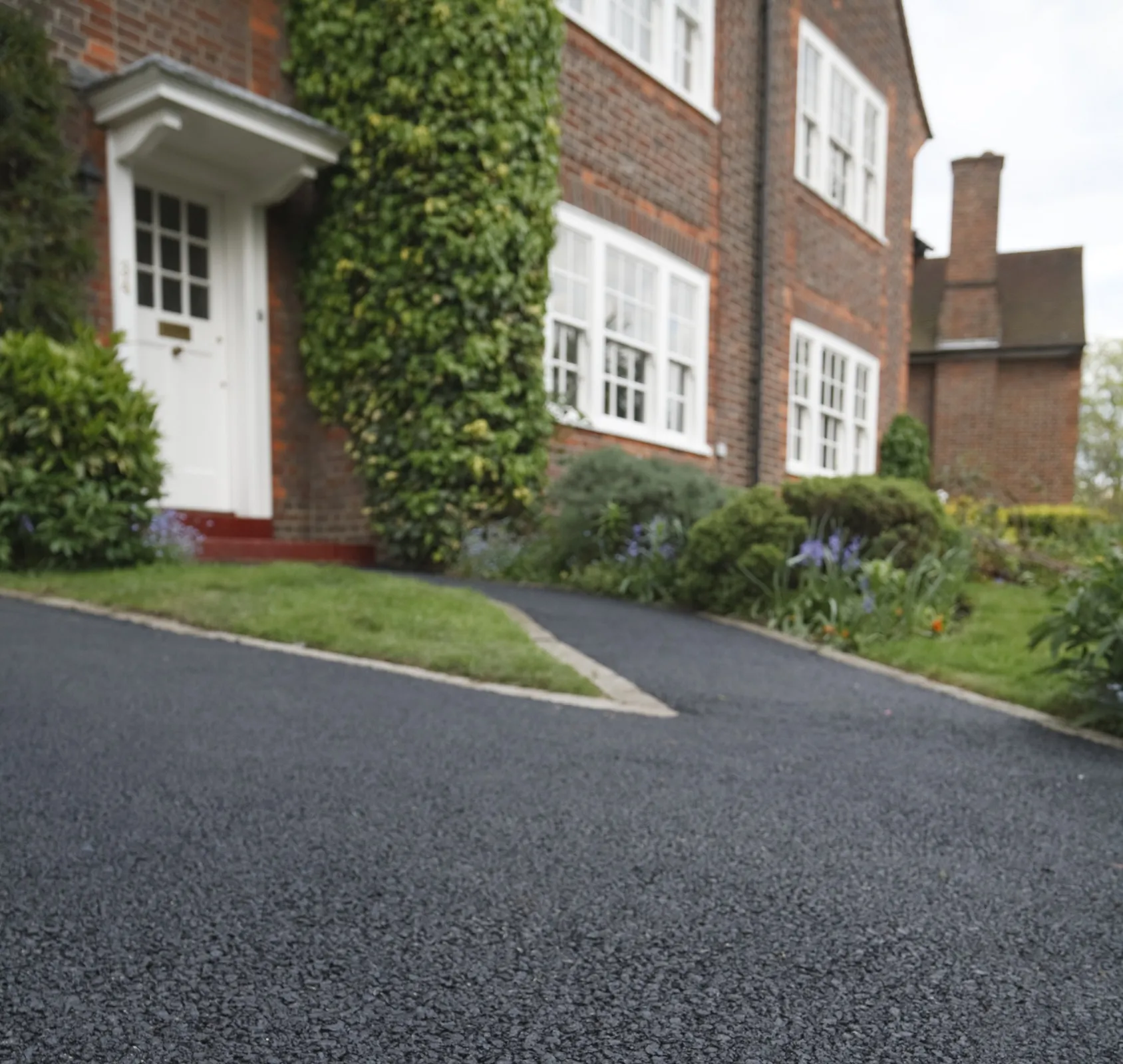 asphalt-b3 Brick house with white windows, a white door, and a newly paved driveway lined with green plants.