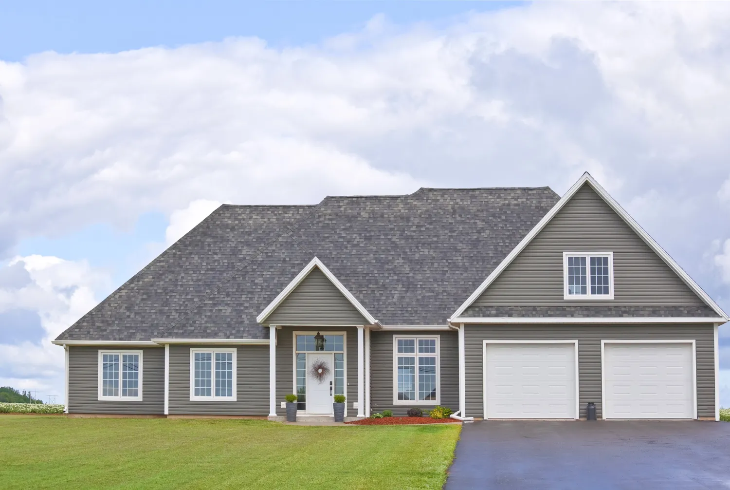 asphalt-home-b Gray suburban house with a large lawn, two-car garage, and cloudy sky in the background.