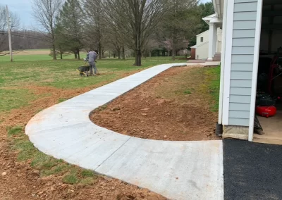 A person uses a wheelbarrow near a freshly poured curved concrete walkway by a house.