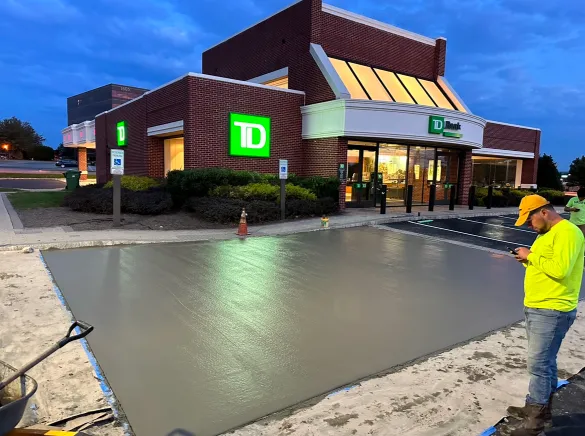 Freshly poured concrete in front of a TD Bank building at dusk; worker in yellow shirt checks phone.