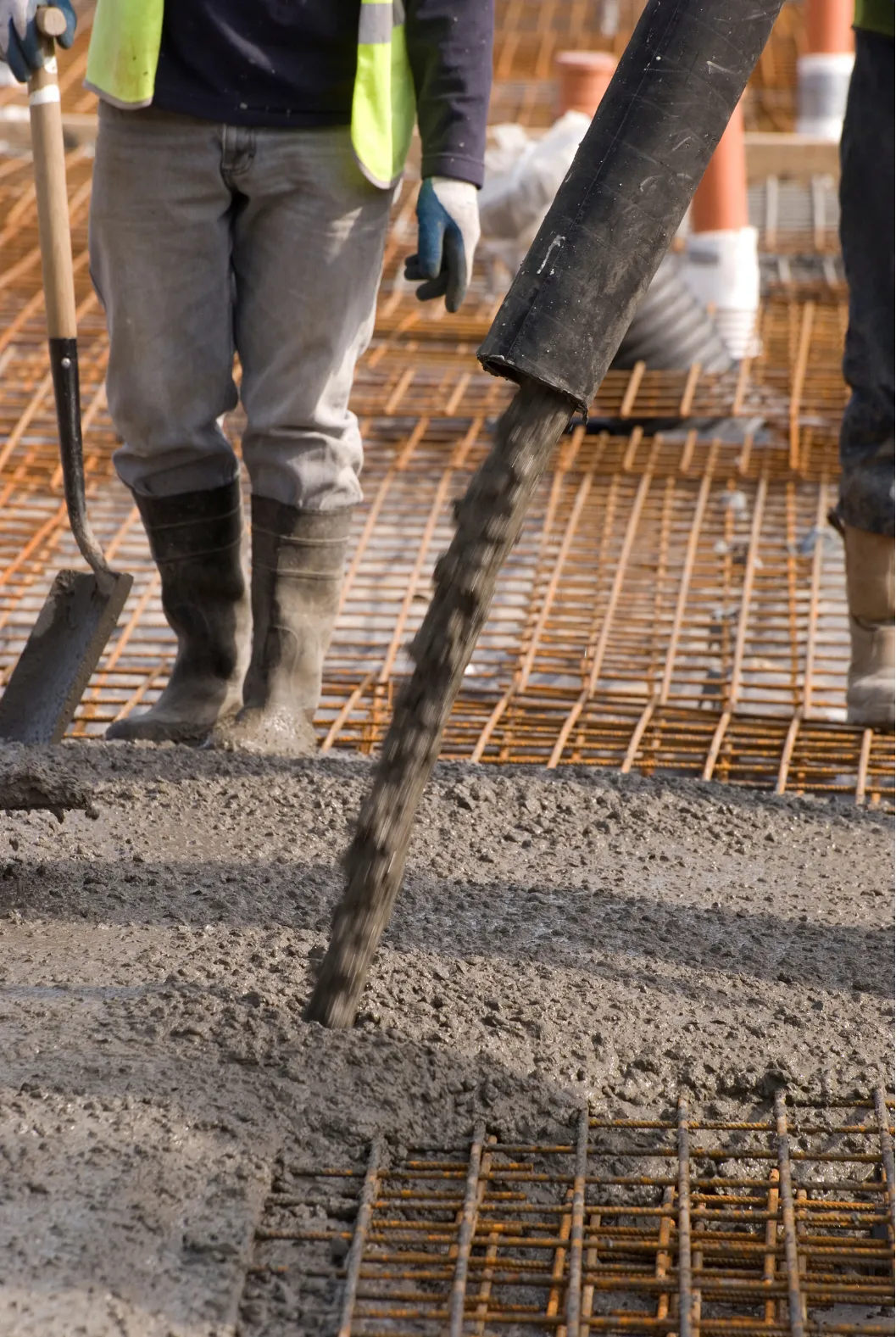 Worker pours wet concrete onto a rebar grid at a construction site, wearing boots and safety vest.