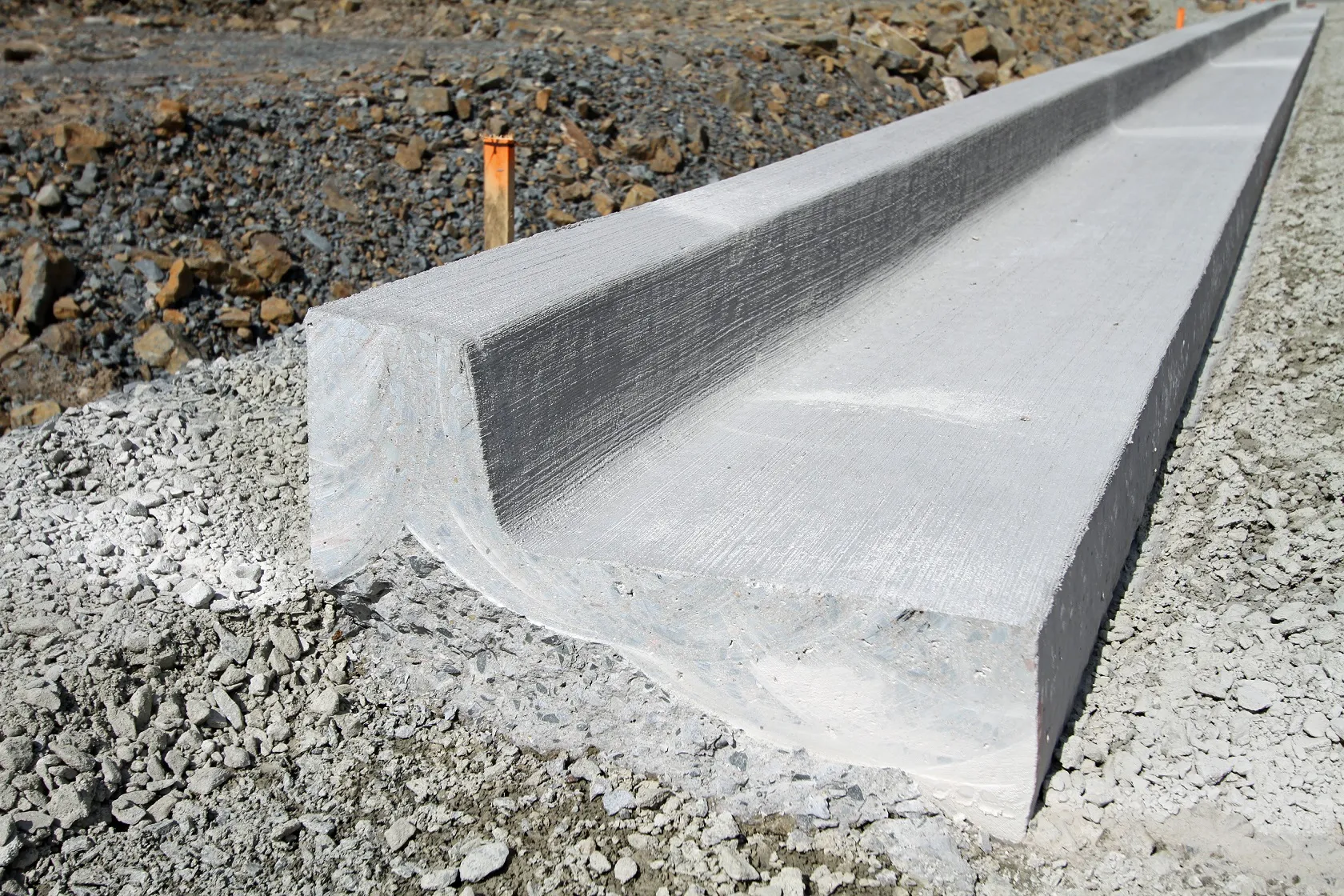 curb-b Concrete drainage channel installation at a construction site, with rough gravel surrounding the channel.