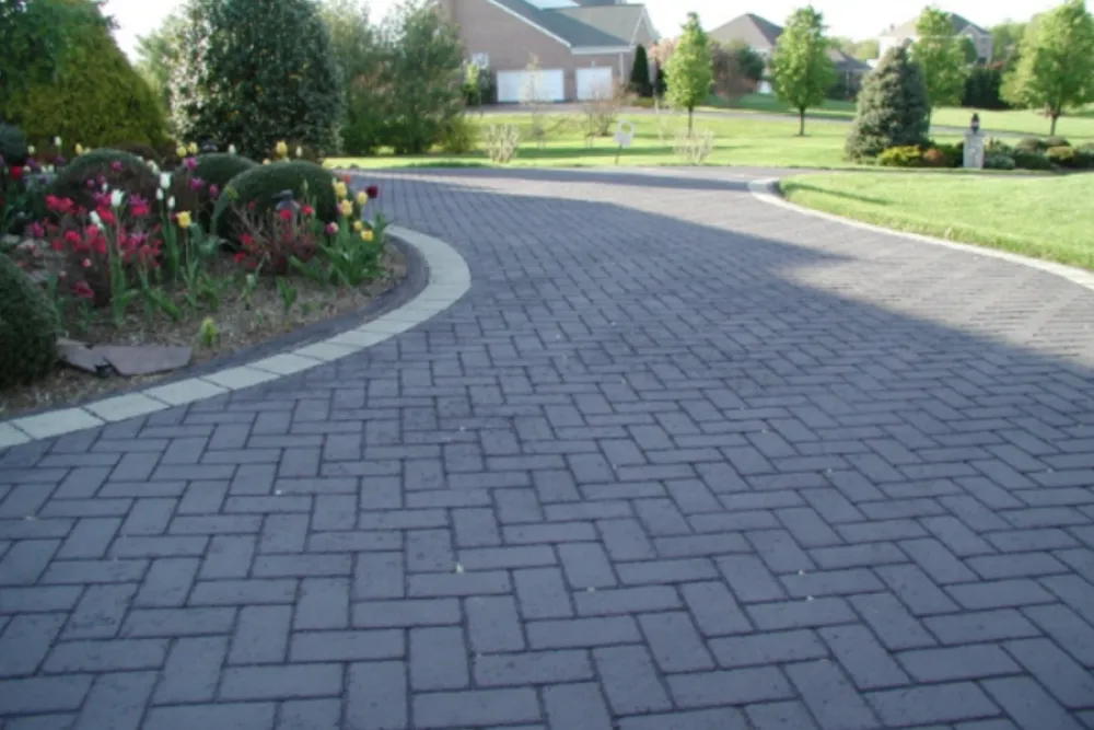 Curved brick driveway with a garden bed of flowers on the left, leading to houses in the background.