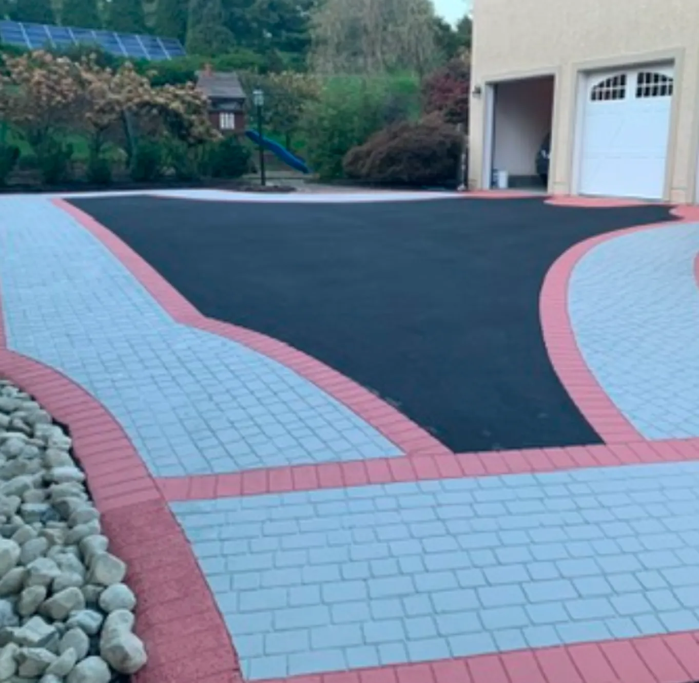 Driveway with blacktop center, light gray stone patterns, pink borders, rocks left, garage door right.