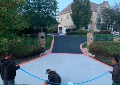 Three people work near a blue rope on a driveway leading to a large house with a blacktop and brick columns.