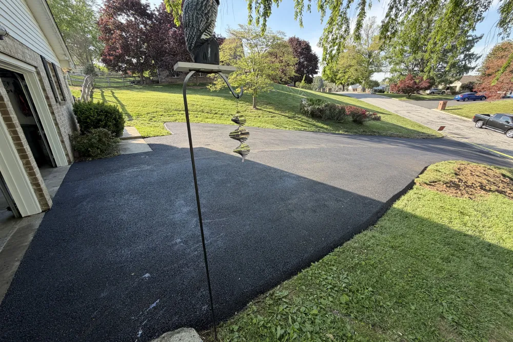 Freshly paved black asphalt driveway beside a garage, bordered by green grass and suburban trees.