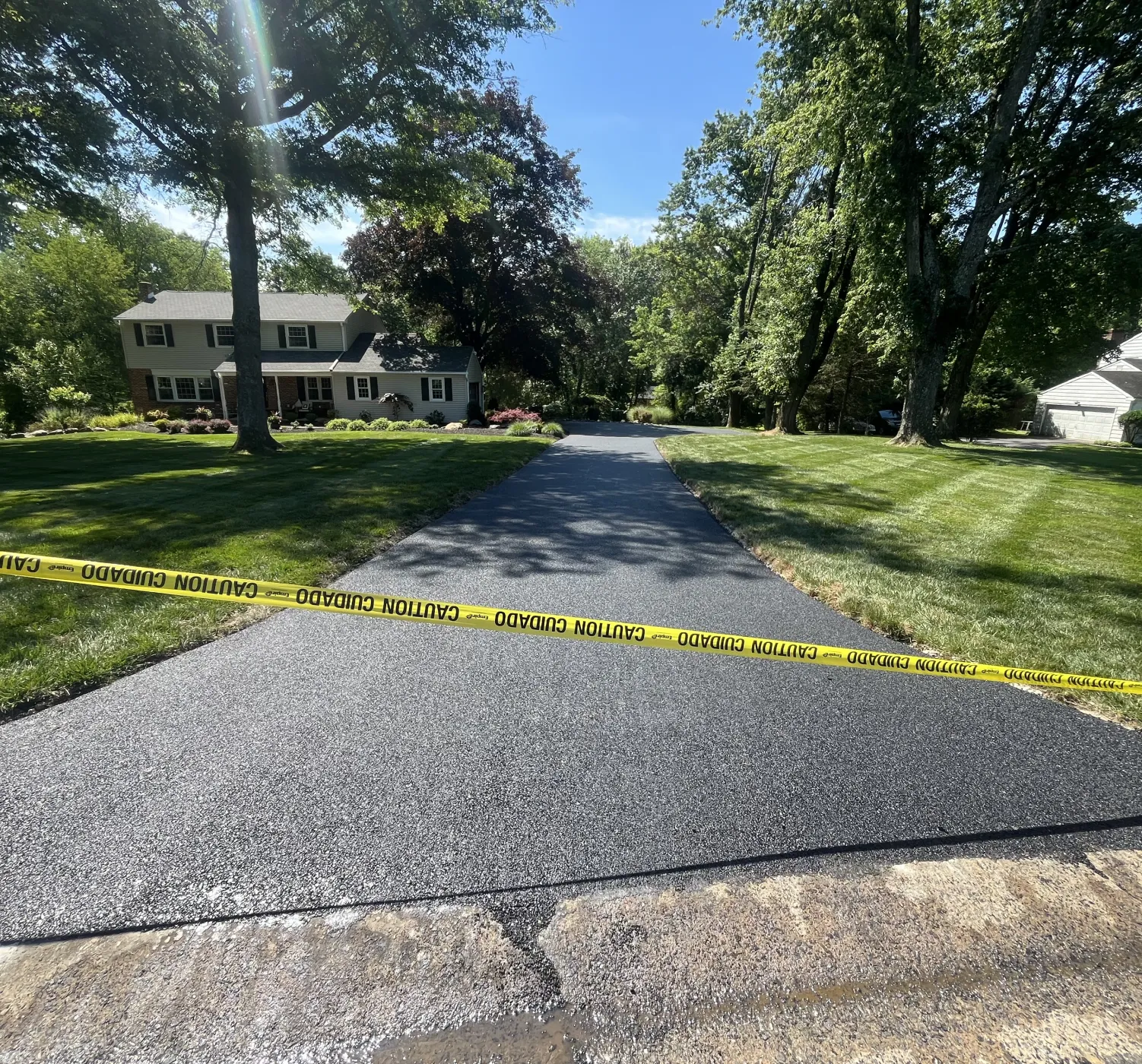 Freshly paved driveway with yellow caution tape in front of a house surrounded by trees and grass.