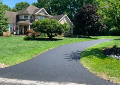 A newly paved black asphalt driveway curves toward a suburban house with a green lawn and trees.