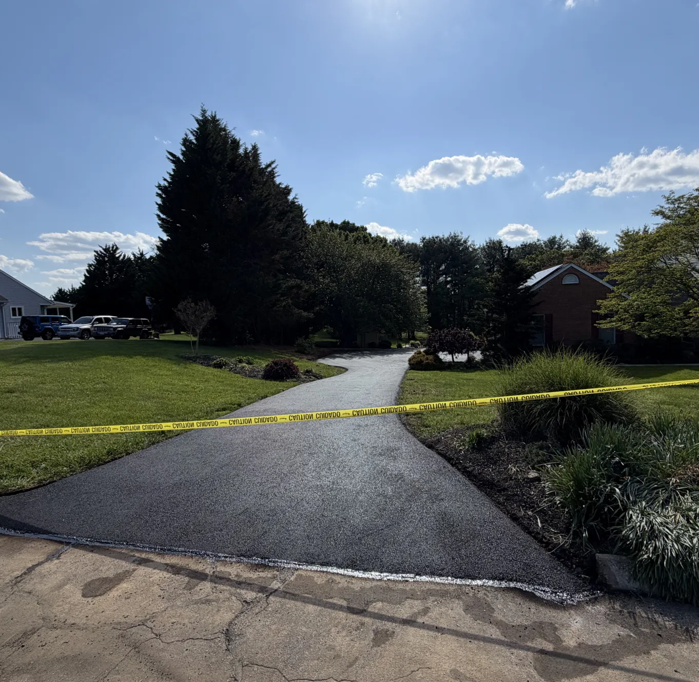 Yellow caution tape blocks access to a freshly paved driveway on a sunny day with scattered clouds.