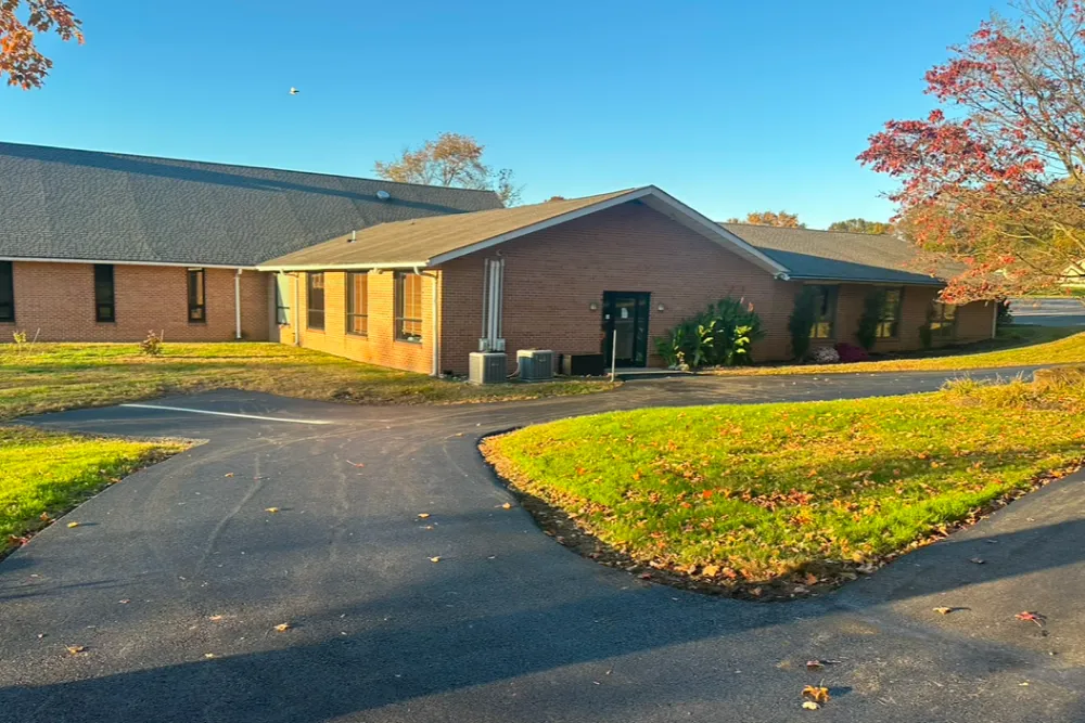 A small brick building with a black door and curved driveway, surrounded by grass and autumn trees.