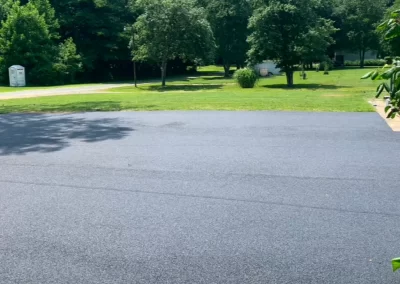 A newly paved asphalt driveway in front of a green grassy yard with trees and a portable restroom in the distance.