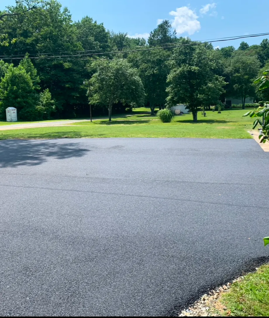 Freshly paved black asphalt driveway bordered by green grass, trees, and a sunny sky in the background.