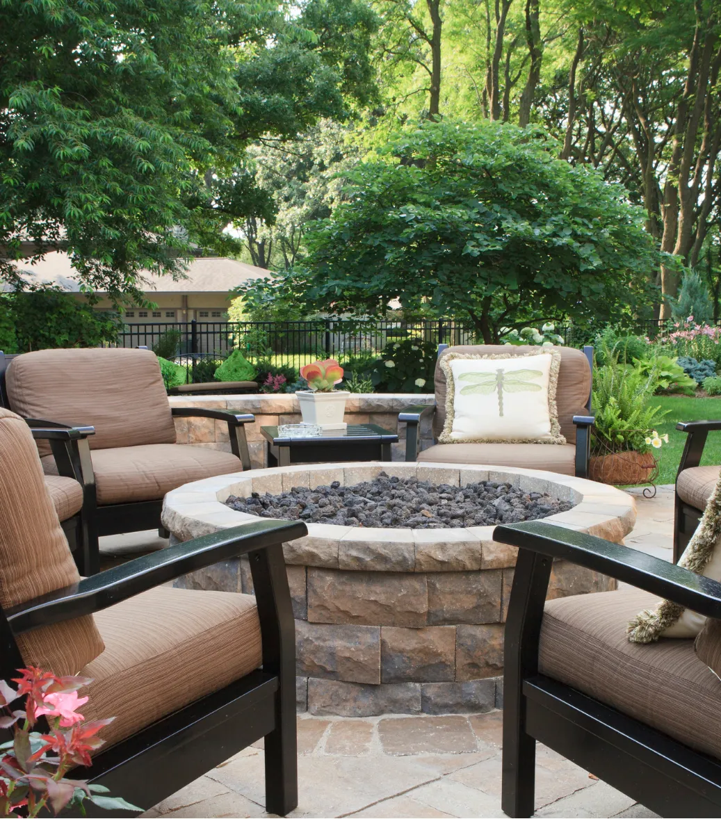 Six cushioned chairs surround a stone firepit on a patio, with lush greenery and trees in the background.