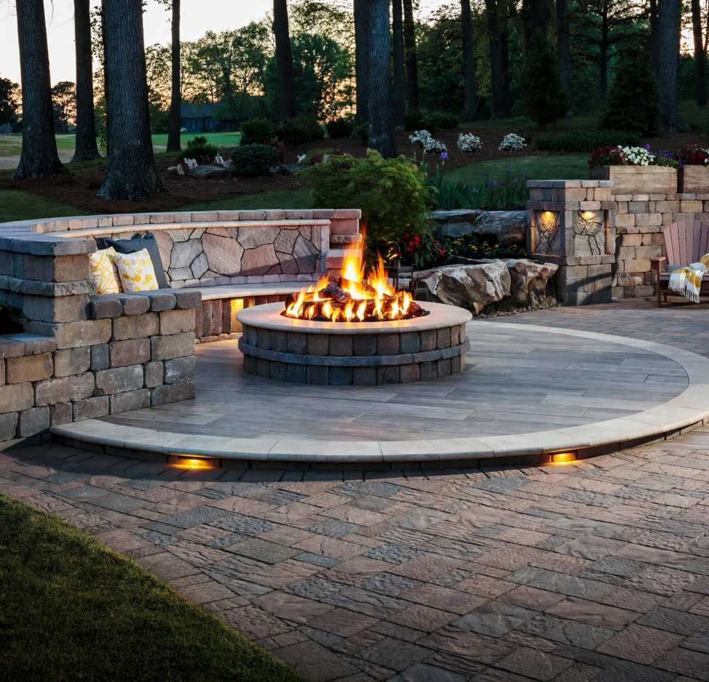 Stone patio with circular fire pit, built-in bench, and soft lighting, surrounded by trees at dusk.