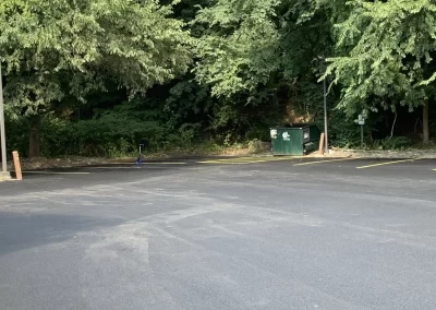 Empty parking lot with a single trash dumpster near a wooded area and tall green trees in the background.