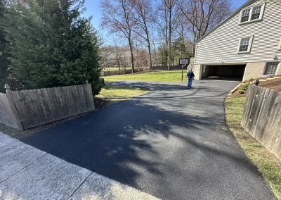 A paved driveway leads to a house with an open garage; a person stands near the basketball hoop.