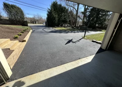 View from a garage looking out at a freshly paved black driveway with a basketball hoop and wooden landscaping stairs.