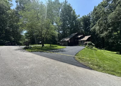 A house sits among trees with a newly paved driveway, cordoned off by yellow tape, on a sunny day.