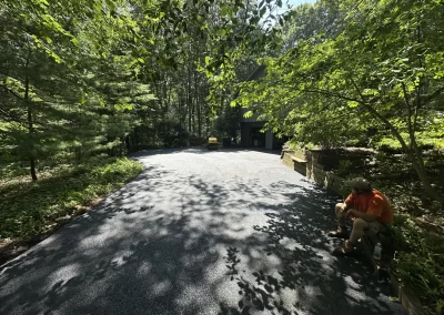 A man sits on the edge of a newly paved driveway in a wooded area with a small steamroller in the background.