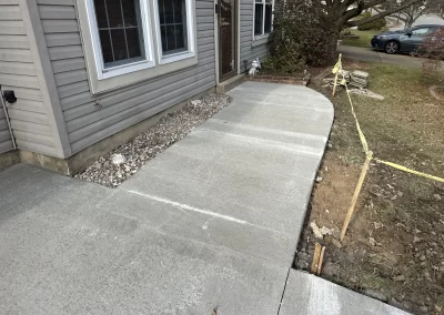 Freshly poured concrete walkway curves to a house entrance; caution tape blocks off unfinished edge.