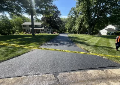 Freshly paved driveway with caution tape blocking access, surrounded by green lawns and trees.