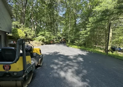 Yellow steamroller on fresh asphalt driveway, surrounded by trees, with workers visible in the background.