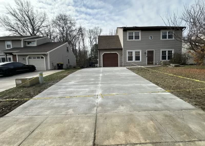 A new concrete driveway with caution tape is in front of a two-story suburban house on a cloudy day.