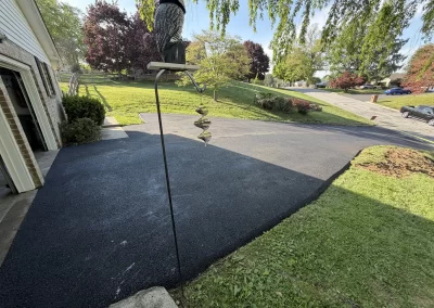 Freshly paved black asphalt driveway next to a green lawn, garage, and tree-lined suburban street.