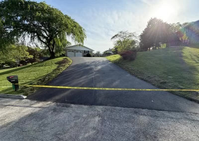 Blacktop driveway blocked by yellow caution tape, leading to a house with sun shining from the right.
