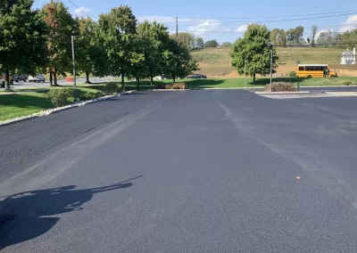 Freshly paved black asphalt parking lot with trees, grass, and a yellow school bus in the background.