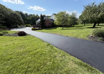 A paved driveway leads to a brick house surrounded by trees and a green lawn under a blue sky.