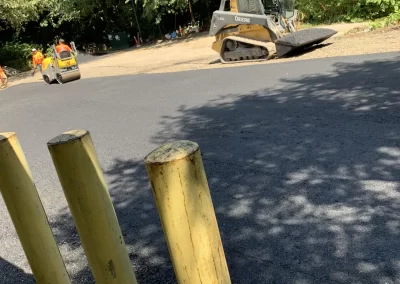 Construction machinery, including a Deere skid steer and a steamroller, paves a fresh asphalt lot near trees.