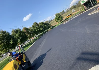 Worker operates steamroller on fresh black asphalt in sunny parking lot near green trees and a school bus.