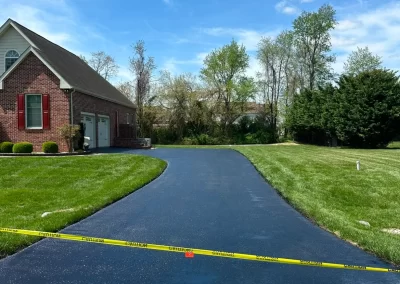 Freshly paved driveway with caution tape, next to a brick house and green lawn under a blue sky.