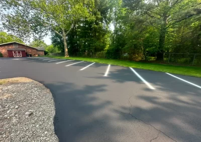 Freshly paved parking lot with white lines, bordered by grass, trees, and a red barn-style building.
