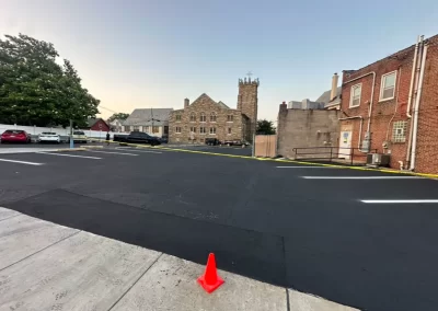 A single orange cone sits on fresh blacktop in a parking lot near brick and stone buildings.