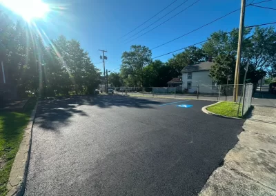 Freshly paved parking lot with one marked accessible spot, trees, and a house in the background on a sunny day.