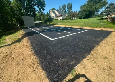 Freshly paved blacktop tennis court with white lines, surrounded by grass and trees, under a clear sky.