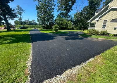 Freshly paved black asphalt driveway curves near a yellow house with green grass and trees around.