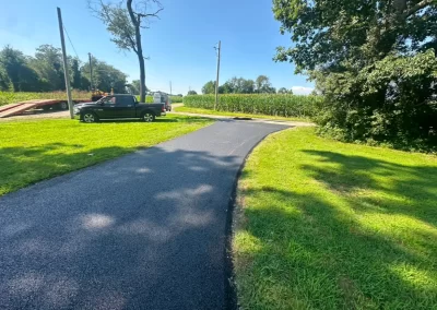 Paved path curving through green grass, with a black truck parked nearby and trees under a blue sky.