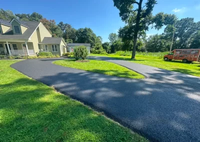 Freshly paved curved driveway next to a house, with green grass and trees, under a clear blue sky.