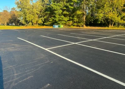 Empty parking lot with fresh pavement and white lines, surrounded by trees and a green dumpster in the back.